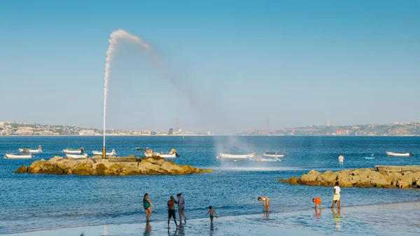 Gente en una playa cerca de Lisboa con un géiser con vistas al icónico puente del 25 de abril y la estatua de Rei Cristo — Foto de Stock