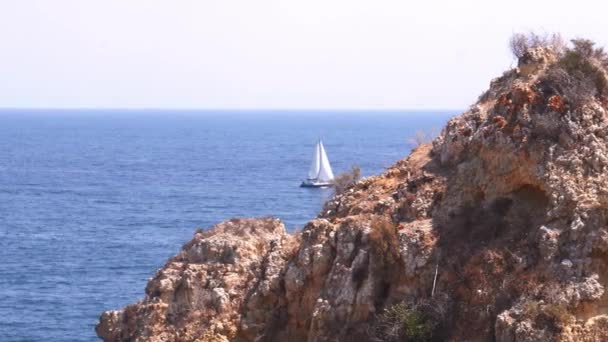 Вітрильний корабель яхта з білими вітрилами у відкритому Атлантичному океані ховається в напрямку скелі в Лагосі, Алгарве, Португалія — стокове відео