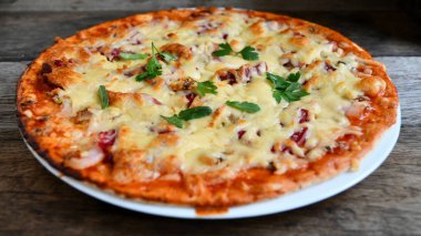 Ahşap bir masa üzerinde beyaz bir seramik tabakta peynir, domates, pastırma ve otlar ile ev yapımı pizza.