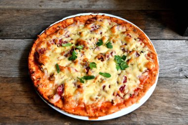 Ahşap bir masa üzerinde beyaz bir seramik tabakta peynir, domates, pastırma ve otlar ile ev yapımı pizza.