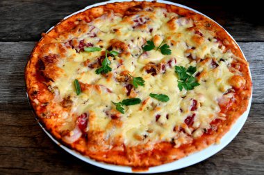 Ahşap bir masa üzerinde beyaz bir seramik tabakta peynir, domates, pastırma ve otlar ile ev yapımı pizza.