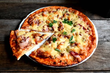 Ahşap bir masa üzerinde beyaz bir seramik tabakta peynir, domates, pastırma ve otlar ile ev yapımı pizza.