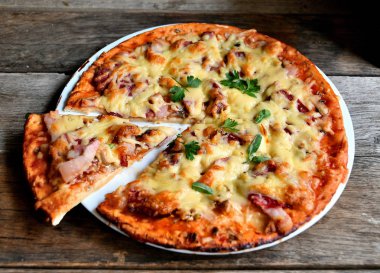 Ahşap bir masa üzerinde beyaz bir seramik tabakta peynir, domates, pastırma ve otlar ile ev yapımı pizza.