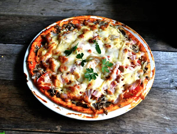 Ahşap bir masa üzerinde beyaz bir seramik tabakta peynir, domates, pastırma ve otlar ile ev yapımı pizza.