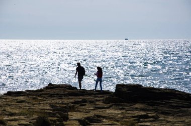 Bir kız ve bir adam deniz kenarında bir kayanın üzerinde yürüyorlar..