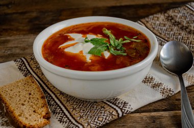 Beyaz bir kasede çorba, ekmek, metal kaşık, mutfaktakiler. Peçete ev mutfağında, ahşap bir masanın üzerinde..