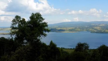 Ağaçların tepesinde bir dağ gölü manzarası. Güneşli bir yaz gününde, mavi gökyüzü ve kabarık bulutlarla insansız hava aracı görüntüsü.