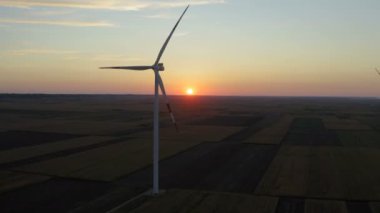 Arkasında güzel bir günbatımı manzarası olan büyük bir tarlanın ortasında büyük bir rüzgar türbininin yanında İHA ile uçmak.