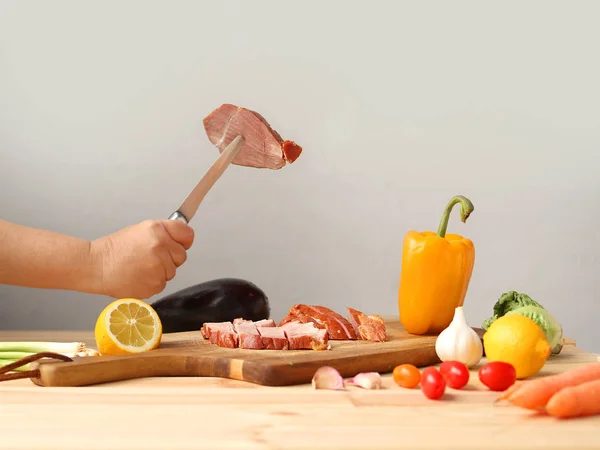 Жінка розрізає м'ясо на дошці, поруч зі свіжими овочами лежать: сирі баклажани, морква, салат, паприка, лимони, зелень крупним планом на світлому фоні — стокове фото