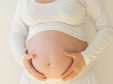 Hamile kadın ışık kapalı evde evde odada beyaz zemin üzerinde bir pencere yanında iki elleri göbek altında tutarak uzun saçlı. Yakın çekim.