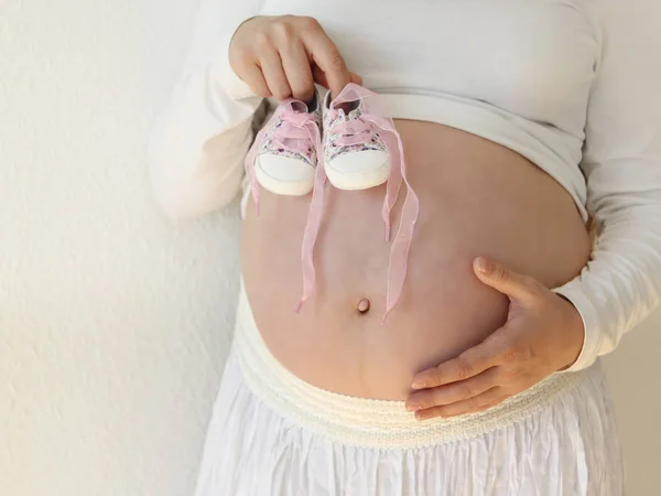 Donna incinta con i capelli lunghi in jeans su uno sfondo bianco tiene nelle mani vicino alla pancia stivaletti bambino nella stanza a casa. La luce proviene da una finestra su uno sfondo bianco carta da parati. Primo piano . — Foto Stock