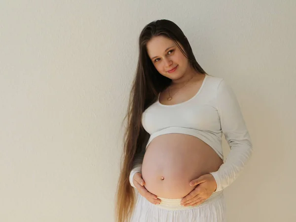 Zwangere vrouw in witte kleren op een witte achtergrond ondersteunt de maag met open handen zachtjes op de kamer thuis. Lampje licht op een witte achtergrond van een venster. Close-up. — Stockfoto