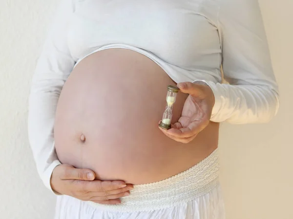 Donna incinta con i capelli lunghi in abiti bianchi su uno sfondo bianco che tiene una clessidra accanto alla pancia nella stanza a casa. La luce proviene da una finestra su uno sfondo bianco carta da parati. Primo piano . — Foto Stock