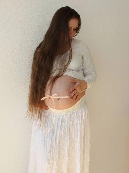 Mulher grávida com cabelos longos em roupas brancas em um fundo branco em um quarto em casa. Barriga amarrada com uma fita de seda com um arco. A luz vem de uma janela em um fundo branco papel de parede. Close-up . — Fotografia de Stock