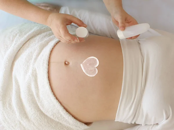 Segurando as mãos de uma jovem mulher abriu um frasco de creme branco e desenha um coração de creme branco no estômago nu de uma mulher grávida deitada. Cuide do seu filho . — Fotografia de Stock