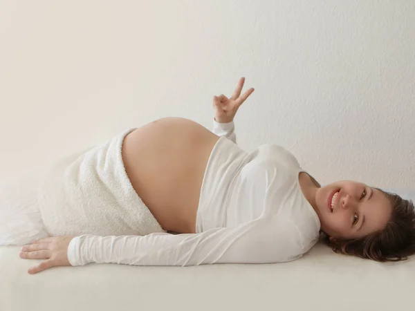 Barriga grande de uma mulher grávida que se deita com roupas brancas nas costas. A mão no fundo branco mostra uma cabra. Close-up . — Fotografia de Stock