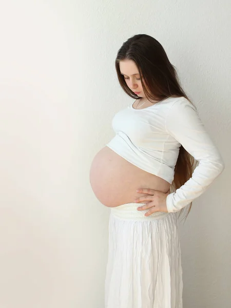 Kobieta w ciąży w białe szaty na białym tle delikatnie obsługuje żołądka z otwartymi rękami w pokoju w domu. Światło pochodzi z okna na tle tapety. Szczelnie-do góry. — Zdjęcie stockowe