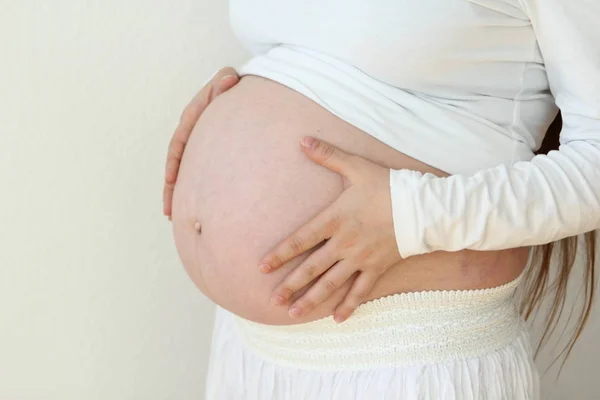 Femeia gravidă cu păr lung, ținându-și cele două mâini sub burtă, ia o poziție laterală lângă o fereastră de lumină în interior, pe un fundal alb, în camera de acasă. Close-up . — Fotografie, imagine de stoc