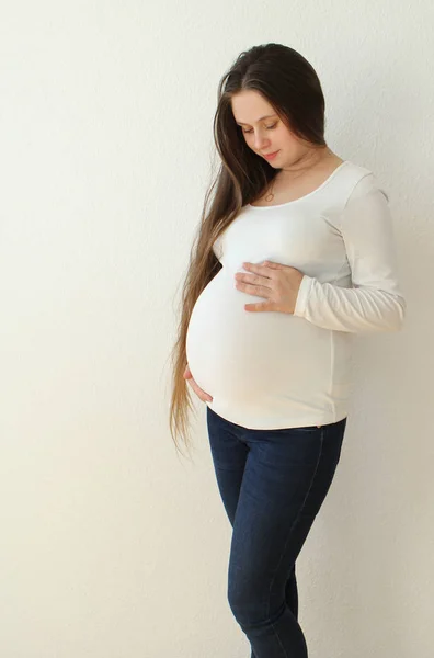 O tânără frumoasă femeie însărcinată în blugi pe un fundal alb își susține ușor stomacul cu mâinile deschise în cameră acasă. Lumina vine de la o fereastră de pe un tapet alb de fundal. Close-up . — Fotografie, imagine de stoc