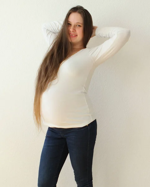 Junge schöne schwangere Frau mit langen Haaren in Jeans auf weißem Hintergrund, die Hände hinter dem Kopf im heimischen Zimmer. Licht kommt aus einem Fenster auf einer weißen Hintergrundtapete. Nahaufnahme. — Stockfoto
