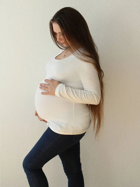 Jovem mulher grávida bonita em jeans em um fundo branco suavemente suporta o estômago com as mãos abertas no quarto em casa. A luz vem de uma janela em um fundo branco papel de parede. Close-up . — Fotografia de Stock