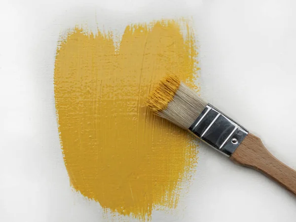 Ein kleiner Pinsel mit gelber Farbe liegt mit frischen gelben Farbstrichen auf einer weißen Fläche. — Stockfoto