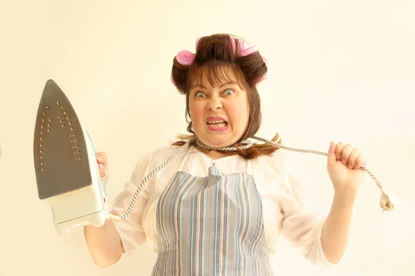 A woman with an iron cord around her neck in home clothes, in an apron, with hair curlers in her hair holding an iron. Portrait close-up