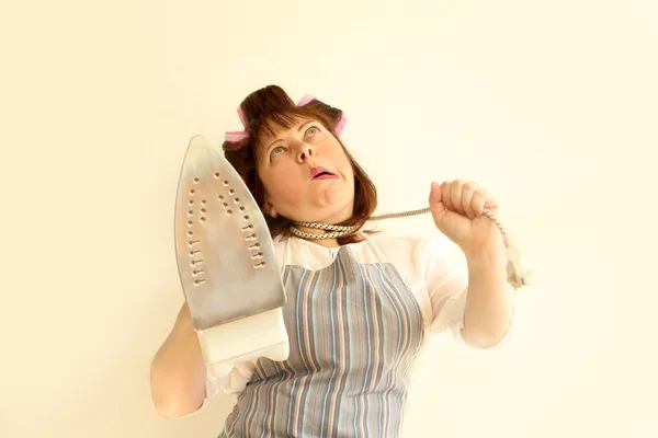 Μια γυναίκα με ένα σιδερένιο καλώδιο γύρω από το λαιμό της με ρούχα στο σπίτι, σε μια ποδιά, με σιδερένιες τρίχες στα μαλλιά της κρατώντας ένα σίδερο. Πορτραίτο κοντινό — Φωτογραφία Αρχείου