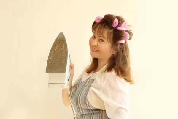 Mulher posando em roupas de casa, em um avental, com encrespadores de cabelo em seu cabelo segurando um ferro. Retrato close-up — Fotografia de Stock