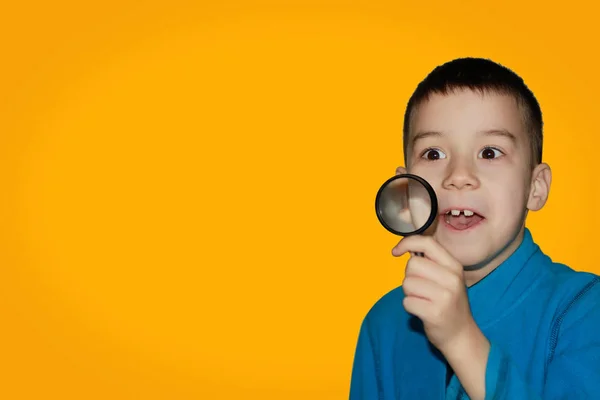 Surprised child looking through a magnifying glass, opening his mouth on a yellow background, close-up — Stock Photo, Image
