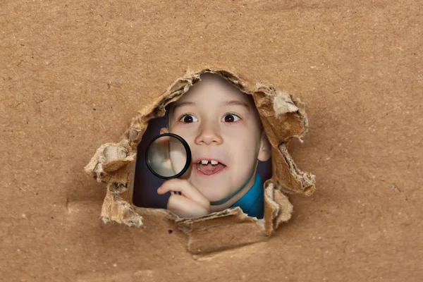 Verbaasd kind kijken door een vergrootglas, opent zijn mond, kijken uit een gat in het karton — Stockfoto