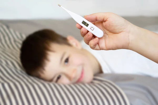 Female hand holds white thermometer on a blurred background with a lying boy, light background — Stock Photo, Image