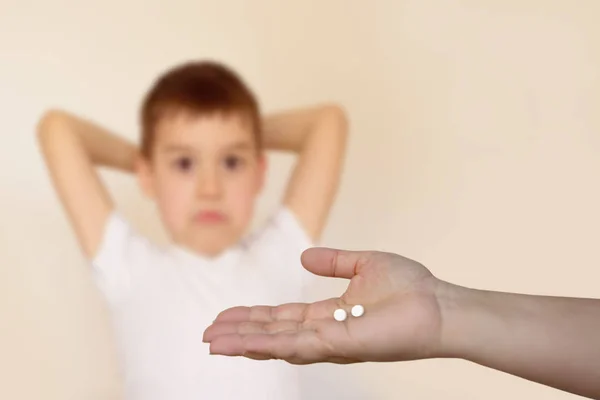 Mano femenina extiende dos píldoras blancas en la palma de la mano sobre un fondo borroso de un niño que levantó las manos detrás de la cabeza con una cara angustiada, fondo claro — Foto de Stock