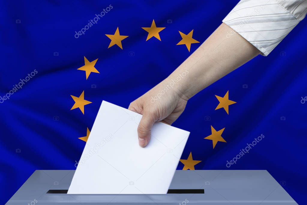 female hand in a white blouse drops a ballot paper in a ballot box against the background of the European Union flag and modern architecture, the concept of elections to the European Parliament