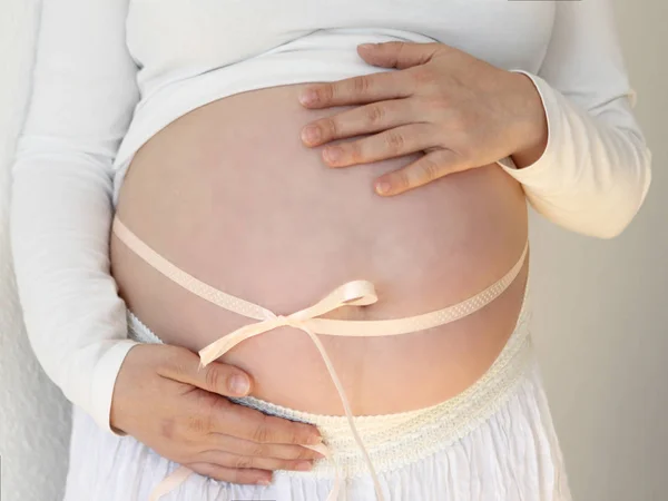 Fată însărcinată cu o burtă mare, legată cu o panglică ușoară, în haine albe, își susține burta cu atenție — Fotografie, imagine de stoc
