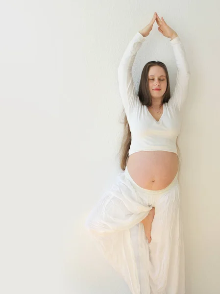 Menina grávida com uma barriga grande, em roupas brancas está de pé, braços levantados, em uma pose de ioga, vertical — Fotografia de Stock