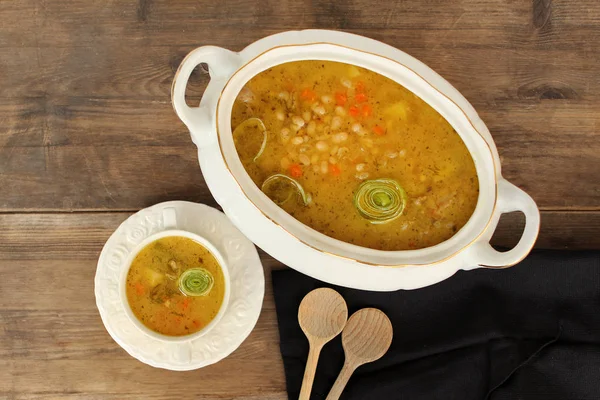 Top view on bean soup in a white plate and white tureen on an old wooden table, billowy black napkin — Stock Photo, Image