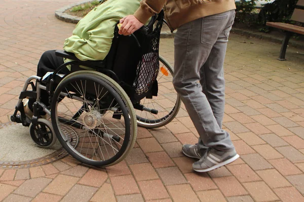 Gri kot pantolonlu bir adam yaşlı bir kadını, sokakta tekerlekli sandalyede engelli bir kişiyi yürüyüşe taşır.