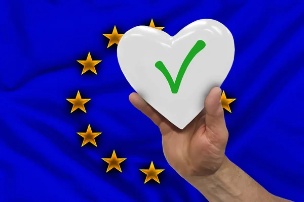 A kéz fehér szívvel rendelkezik, az uniós zászló hátterével szemben az Európai Parlament választásszimbólumai a hang jele. — Stock Fotó