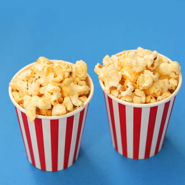 Palomitas crujientes aromatizadas vertidas en dos tazas de papel blanco y rojo a rayas sobre un fondo azul, espacio para copiar, primer plano, cuadrado — Foto de Stock