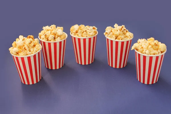 Palomitas de maíz crujientes aromatizadas se vierten en cinco tazas de papel blanco y rojo rayado que se colocan sobre un fondo lila semi-aceitado, espacio de copia en el centro — Foto de Stock