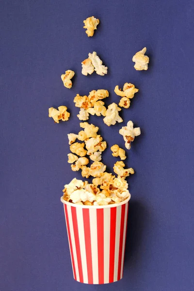 Palomitas crujientes saborizadas goteando desde arriba en una taza de papel blanco-rojo rayado sobre un fondo lila, espacio de copia — Foto de Stock