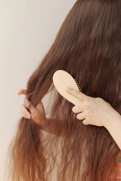 Menina com cabelos castanhos compridos penteando-os com um pente de madeira, segurando fios, vertical, close-up — Fotografia de Stock