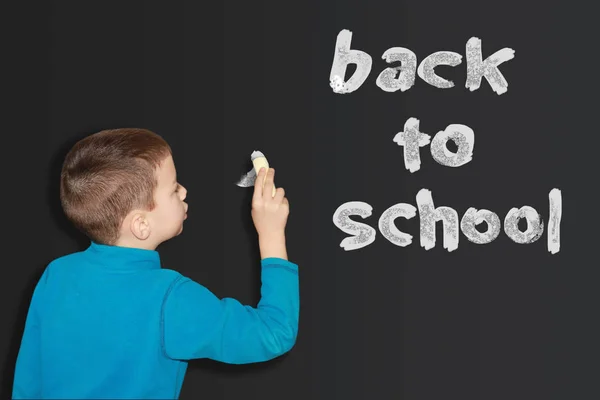 Rapaz de camisola azul escreve num quadro negro com giz branco, vista traseira. Voltar ao conceito de escola — Fotografia de Stock