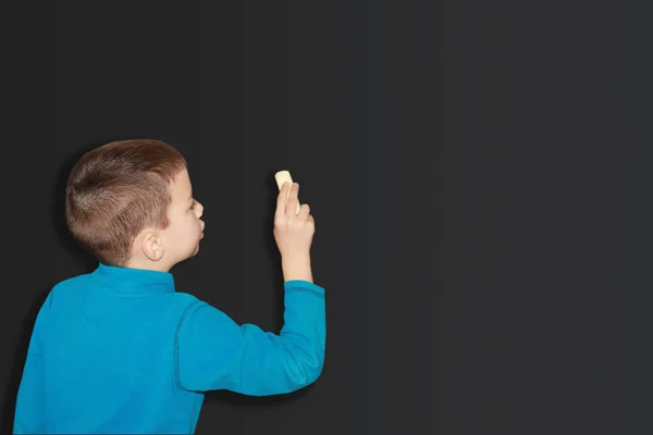 Chłopiec w niebieskim swetrze pisze na czarnej tablicy szkolnej z białą kredą, widok z tyłu — Zdjęcie stockowe