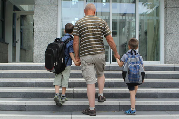 Мужчина ведет мальчиков со школьными сумками, держаться за руки, ходить в школу, вернуться к школьной концепции — стоковое фото