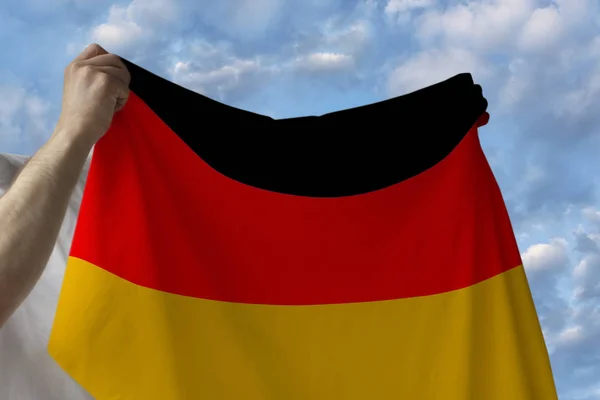 Beautiful national flag of the country of Germany in male hands against the blue sky with clouds — Stock Photo, Image