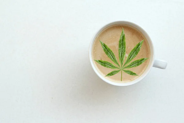 Foglia verde di canapa, marijuana in una tazza larga bianca di caffè, cappuccino con latte, cacao con schiuma di cuore su un tavolo bianco, vista dall'alto, risparmiare spazio, primo piano, concetto — Foto Stock
