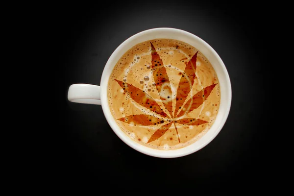 Folha de cannabis, maconha em uma ampla xícara de café branco com crema de coração, cappuccino em um fundo preto com vinheta, vista superior, economizar espaço, close-up — Fotografia de Stock