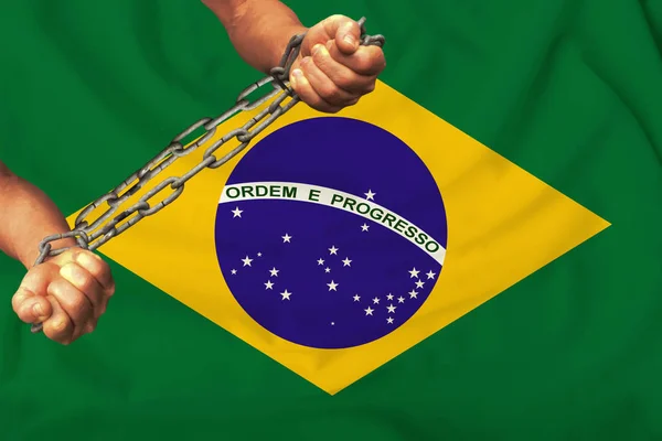 Mãos masculinas acorrentadas em cadeias de ferro pesado contra o fundo da bandeira do Brasil em uma seda suave com dobras no vento, o conceito do movimento pelos direitos humanos — Fotografia de Stock